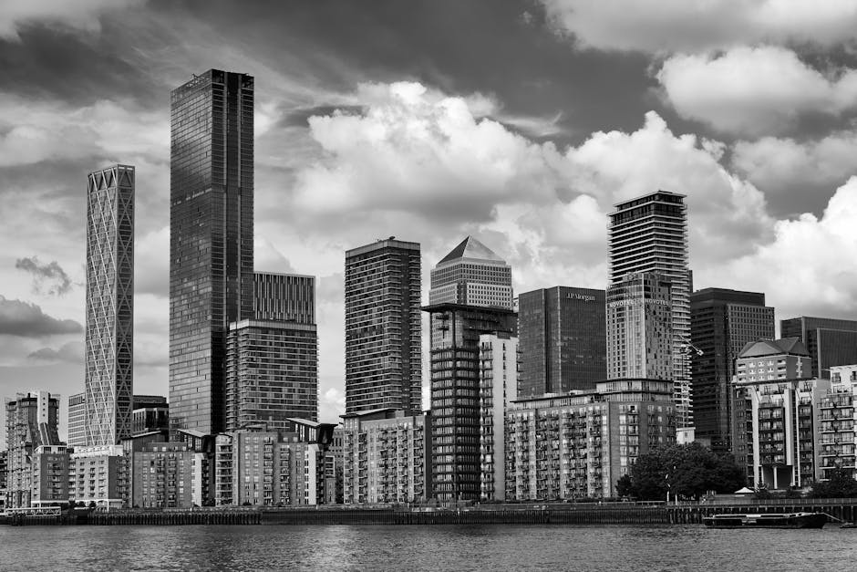 A black and white photograph of a city skyline featuring a variety of modern high-rise buildings, some with reflective glass facades and geometric architectural designs, situated along a waterfront. The buildings include a tall, rectangular tower with a smooth glass exterior, a twisted tower with diagonal structural elements, and several other skyscrapers with different heights and angular outlines, creating a diverse urban silhouette. In the foreground, there is a row of mid-rise residential or commercial buildings with brick or concrete finishes and numerous windows. The scene is illuminated by natural daylight, with a sky filled with large, fluffy clouds. The overall atmosphere reflects an urban environment where private waste management services, such as those provided by Waste Collection Canary Wharf, could facilitate alternative rubbish removal solutions in busy financial districts or residential developments near the waterfront.
