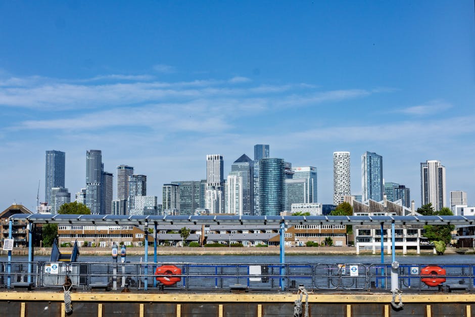 A wide view of the London city skyline taken from the riverbank, featuring a clear blue sky with interspersed wispy clouds. In the foreground, there is a riverside walkway with a metal railing painted in blue, equipped with safety features such as life rings and warning signs. Behind the railing, the river is visible, with a section of a navigation mooring or dock structure that includes metal poles and a horizontal beam detail. Beyond the river, low-rise modern residential buildings with flat roofs, large windows, and exterior finishes in neutral tones are seen, contrasting with the high-rise office skyscrapers and commercial towers that dominate the background. The cityscape includes clustered glass and steel office blocks of varying heights, some with distinct geometric shapes and reflective surfaces that mirror the sky and surrounding structures. The environment suggests an urban waterside area, possibly part of a financial or commercial district, emphasizing the contrast between the natural river setting and the dense concentration of contemporary architecture. This scene aligns with the concept of private or independent waste management solutions, relevant to the surrounding urban infrastructure, when considering the broader context of rubbish removal services in a city environment, as reflected by the domain of wastecollectioncanarywharf.co.uk.