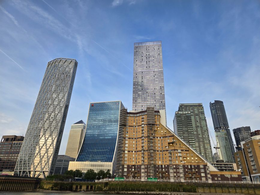A collection of modern high-rise buildings in an urban cityscape viewed from street level during daytime. The image features a tall, rectangular glass skyscraper at the center with a reflective surface, surrounded by other commercial towers with varying architectural styles, including one with a distinctive twisted design on the left, and several with glass facades showcasing different finishes and textures. In the foreground, at the base of the buildings, there are brown brick residential structures with multiple small windows and protruding balconies, situated along a paved road. The sky above is clear with a few wispy clouds and visible contrails, contributing to a crisp, bright atmosphere. This scene captures the dense, diverse architecture typical of central business districts, highlighting the mix of contemporary office buildings and residential units, with subtle references to alternative waste handling environments seen in the context of urban rubbish management, as represented by waste collection services in such contexts, although no waste or disposal activities are visible in the image.