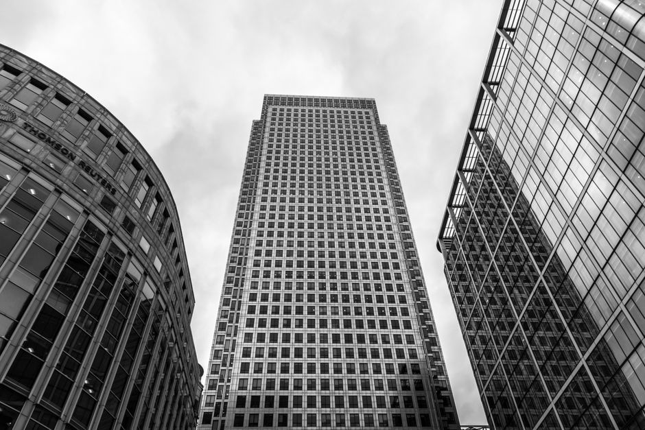 A collection of modern high-rise buildings in an urban cityscape viewed from street level during daytime. The image features a tall, rectangular glass skyscraper at the center with a reflective surface, surrounded by other commercial towers with varying architectural styles, including one with a distinctive twisted design on the left, and several with glass facades showcasing different finishes and textures. In the foreground, at the base of the buildings, there are brown brick residential structures with multiple small windows and protruding balconies, situated along a paved road. The sky above is clear with a few wispy clouds and visible contrails, contributing to a crisp, bright atmosphere. This scene captures the dense, diverse architecture typical of central business districts, highlighting the mix of contemporary office buildings and residential units, with subtle references to alternative waste handling environments seen in the context of urban rubbish management, as represented by waste collection services in such contexts, although no waste or disposal activities are visible in the image.