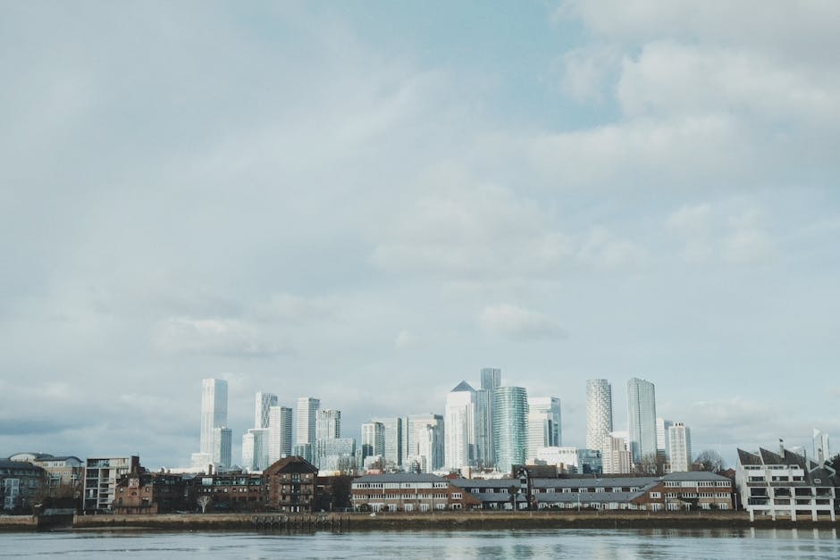 A wide panoramic view of a modern city skyline featuring numerous tall, glass-clad skyscrapers with reflective surfaces, set against a cloudy sky. In the foreground, there is a row of older, low-rise brick buildings and warehouses situated along the water's edge, with some structures featuring white window frames and dark roofs. The river or body of water in the lower part of the image is calm, reflecting parts of the buildings and indicating a tranquil environment. The overall scene suggests a contrasting urban landscape with recent high-rise development behind a historic, operational waterfront area. This urban setting exemplifies the transition from traditional to contemporary architecture, aligning with themes of urban renewal and private waste management services that support efficient rubbish collection within such dynamic environments.