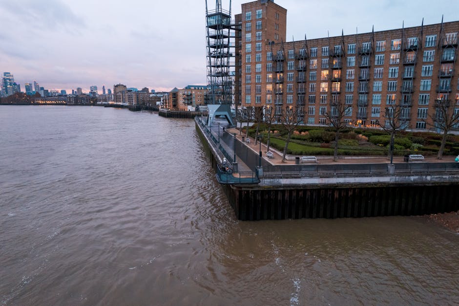 An elevated riverside walkway adjacent to a large, modern multi-storey residential building constructed with red brick and featuring numerous balconies with metal railings. The walkway is bordered by a low concrete wall and has several evenly spaced benches and a few young, leafless trees planted along the edge. A metal staircase with a spiral design provides access to the walkway from the ground level. Beyond the wall, a body of water with a gentle current extends across the foreground, with a distant city skyline visible on the horizon, composed of various high-rise buildings. The sky overhead is overcast, casting a subdued light over the scene, which reflects a typical urban riverside environment suitable for private waste collection or on-site clearance services by waste management specialists such as Waste Collection Canary Wharf, providing efficient rubbish removal in the area.