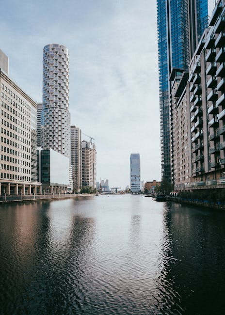 A wide panoramic view of a modern city skyline featuring numerous tall, glass-clad skyscrapers with reflective surfaces, set against a cloudy sky. In the foreground, there is a row of older, low-rise brick buildings and warehouses situated along the water's edge, with some structures featuring white window frames and dark roofs. The river or body of water in the lower part of the image is calm, reflecting parts of the buildings and indicating a tranquil environment. The overall scene suggests a contrasting urban landscape with recent high-rise development behind a historic, operational waterfront area. This urban setting exemplifies the transition from traditional to contemporary architecture, aligning with themes of urban renewal and private waste management services that support efficient rubbish collection within such dynamic environments.