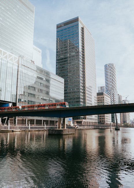 A modern urban scene featuring a river running through a city with tall, contemporary office buildings made of glass and steel in the background. In the foreground, a bridge spans across the river, supporting a bright red train travelling along the elevated tracks. The buildings reflect the sky and surrounding architecture on their glass facades, with some structures displaying a grid-like pattern and others with curved or textured finishes. The water below shows gentle ripples, and the overall atmosphere appears clear with diffuse daylight. This scene illustrates a typical cityscape, which could be associated with private or independent transport or waste collection services operating in metropolitan environments, highlighting the connection between urban infrastructure and service logistics for waste management companies like Waste Collection Canary Wharf in busy financial districts.