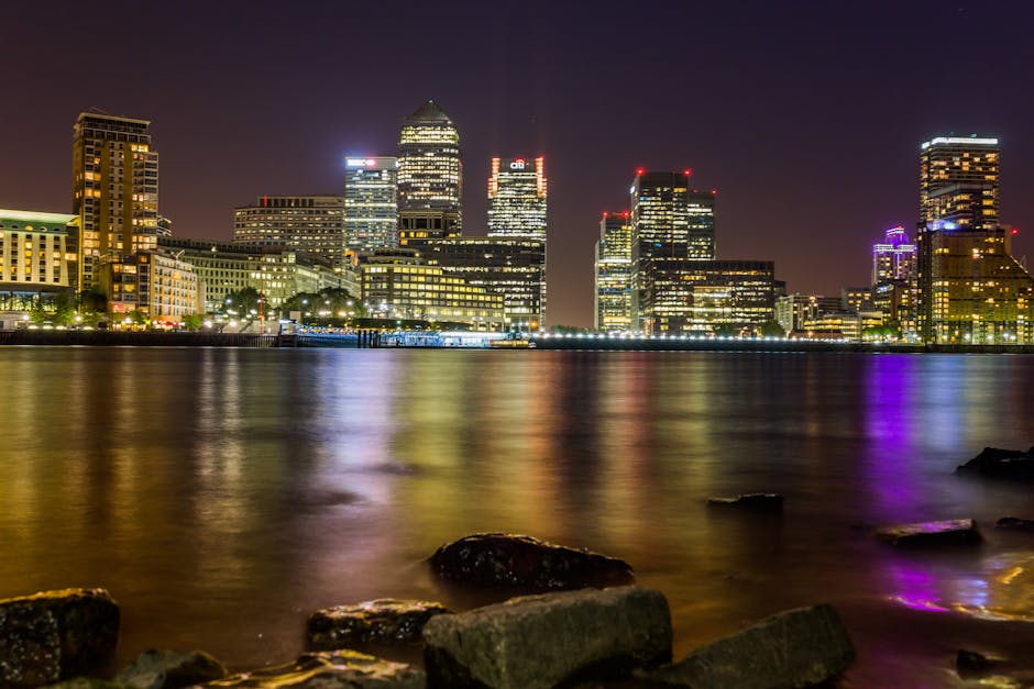 A panoramic view of a modern city skyline featuring numerous high-rise office buildings and skyscrapers made of glass and steel, some under construction with cranes visible atop them. The buildings vary in height and architectural style, with reflective surfaces capturing sunlight. In the foreground, a lush green park area with mature deciduous trees displaying vibrant green foliage stretches across the lower part of the image, creating a natural contrast to the urban landscape. The sky above is partly cloudy with wispy white clouds dispersed across a bright blue background. The scene appears to be taken during daytime under natural lighting, emphasizing the clarity and detail of both the cityscape and the greenery, suggesting an environment where urban development coexists with maintained public parks, reflecting a focus on alternate waste handling and urban management including private rubbish removal services that might operate within or around this city area.