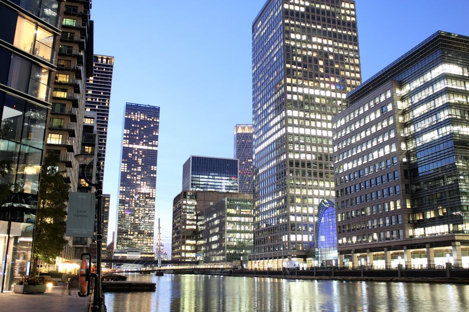 A cityscape featuring a collection of modern high-rise buildings along a waterway during twilight, with the facades of the structures illuminated by interior lights through numerous glass windows. The buildings are made of steel and glass, displaying reflective surfaces and sleek finishes in shades of blue, gray, and black, with some showcasing distinct architectural features such as grid patterns, curvilinear shapes, and varied heights. In the foreground, a promenade with paved walkways, small potted plants, and a lamp post is visible on the left side, suggesting an urban environment suited for pedestrian activity. The calm water reflects the illuminated buildings, creating a mirrored effect that enhances the scene's vibrancy. This urban waterfront scene reflects the typical environment where private waste collection services, such as those offered by Waste Collection Canary Wharf, might operate to manage rubbish clearance from these commercial high-rise complexes, emphasizing the importance of efficient waste handling in densely built-up business districts.